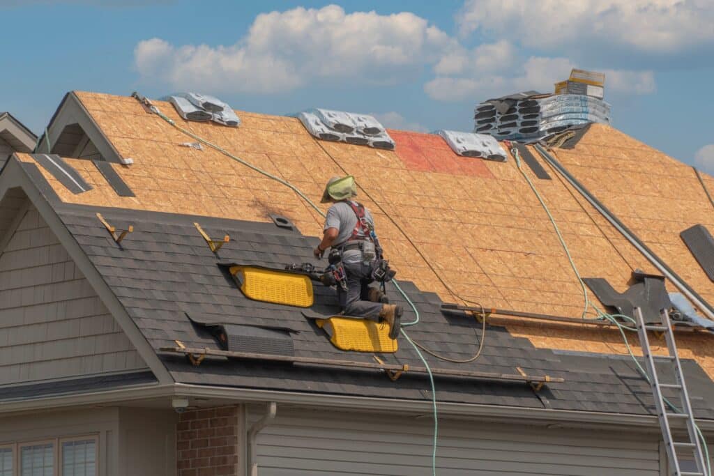 A construction worker wearing safety gear installs shingles during a roof replacement under a blue sky with clouds. The roof is partially finished, with stacks of materials and exposed wooden sections visible.