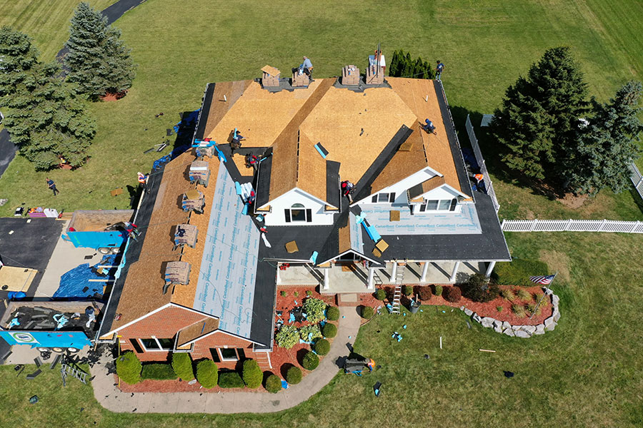 Aerial view of a house undergoing a roof replacement, with workers installing new roofing materials. Some sections reveal exposed wooden sheathing, while others are partially covered with roofing felt and shingles. Lush lawn and trees surround the home.