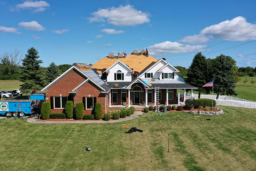 Aerial view of a large two-story house undergoing roof replacement. Workers are replacing the roofing, with wood exposed and equipment scattered on the lawn. A blue truck is parked nearby under a clear sky with a few clouds.