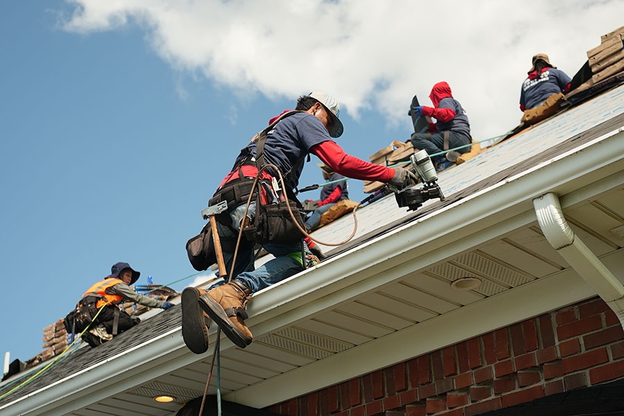 Several workers wearing safety harnesses install shingles on a house roof under a blue sky during a roof replacement, with one worker using a nail gun near the edge and others placing shingles in the background.