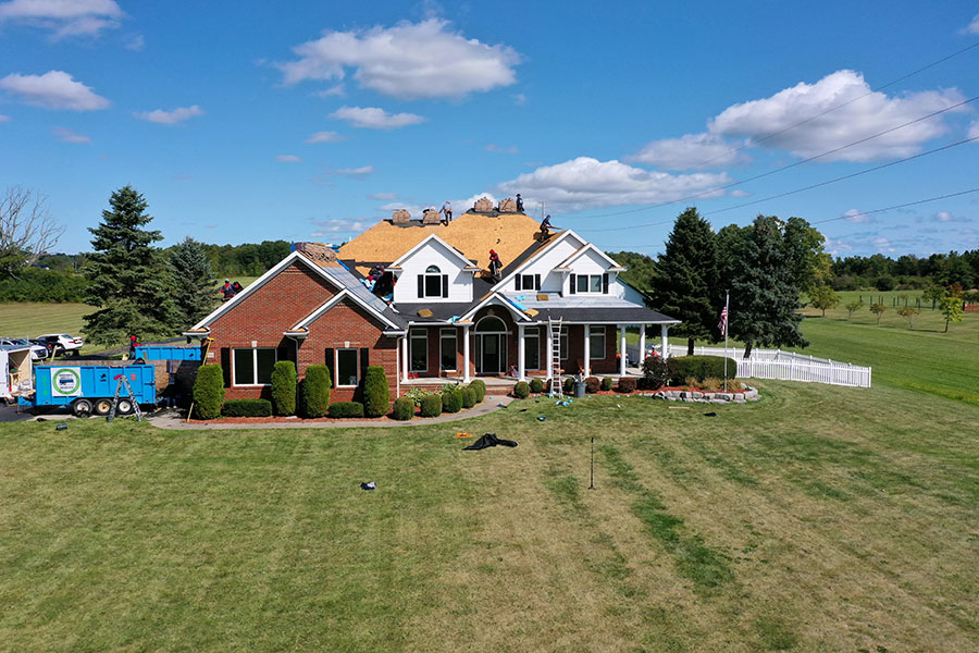 A large suburban house undergoing roof replacement, with workers and construction equipment on site. Some shingles are removed, and a blue truck sits in the yard, surrounded by green grass, trees, a white fence, and a blue sky with clouds.