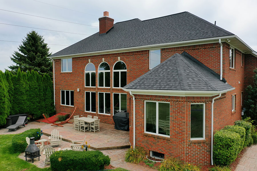 A two-story red brick house with large arched windows, a new roof replacement featuring dark shingles, and a spacious patio with outdoor seating, a dining table, a grill, Adirondack chairs, and lush surrounding greenery.