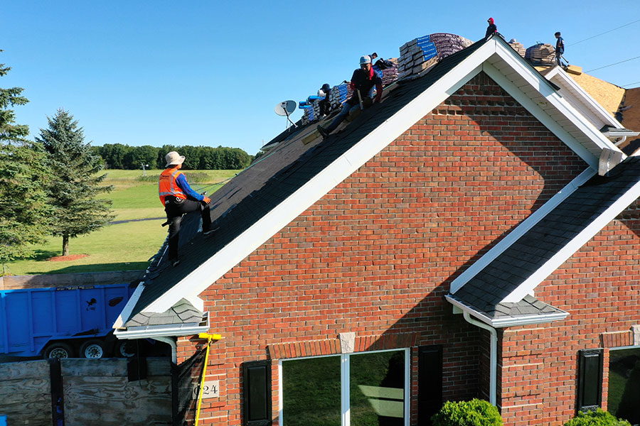Several workers wearing safety gear are completing a roof replacement, installing shingles on the steep roof of a red brick house. Bundles of roofing materials are stacked nearby, with a blue dumpster close to the scene. Trees and an open field are in the background.