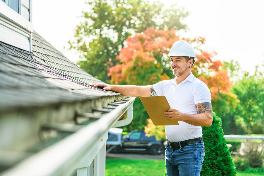 A man wearing a white hard hat and holding a clipboard inspects a house roof and gutter outside on a sunny day, checking for signs that impact roof durability, with green trees and a parked car in the background.
