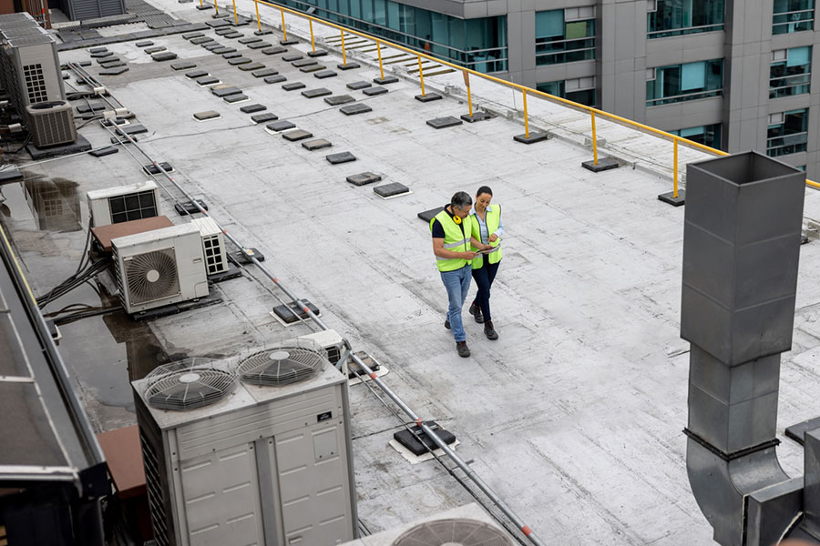 Two people in reflective vests walk on a rooftop with air conditioning units and safety railings, near a modern building—perfectly illustrating the attention to detail essential for commercial roofing projects.
