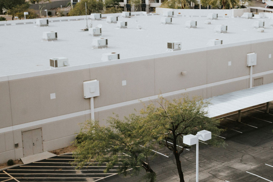 Large beige commercial building with a flat roof featuring numerous HVAC units on top—an ideal example for Michigan business owners seeking insights from a reliable roofing guide. The property also includes empty parking spaces, a few trees, and a metal carport, all highlighting classic commercial roofing details.