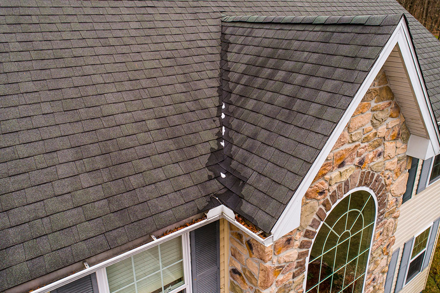 A close-up, angled view of a house roof with dark gray shingles and a stone exterior wall. The roofline displays clear signs of wind damaged roof, with missing shingles along the ridge, and leaves collected in the gutter below.
