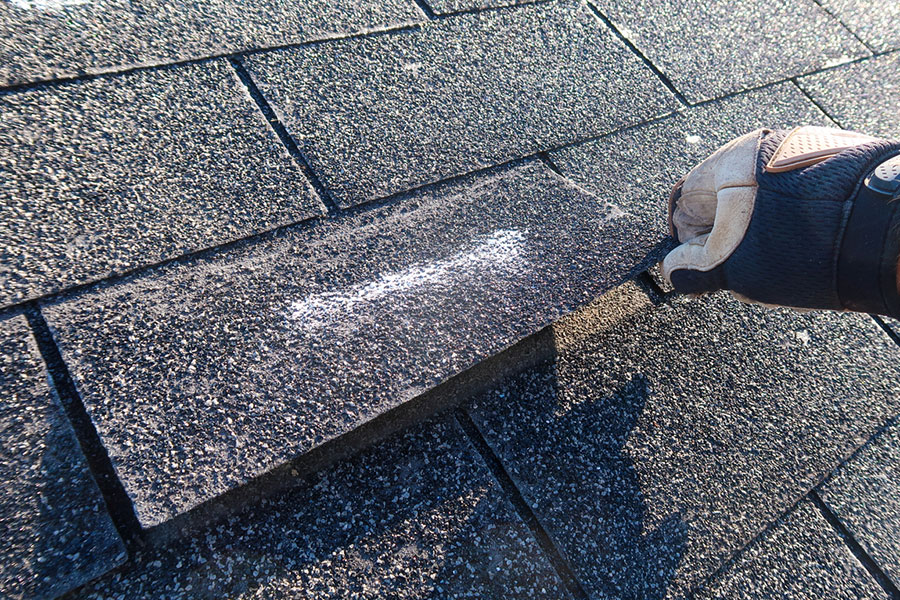A person wearing a work glove lifts a shingle on a residential roof to reveal a white chalk mark, identifying one of the common roof leak causes.